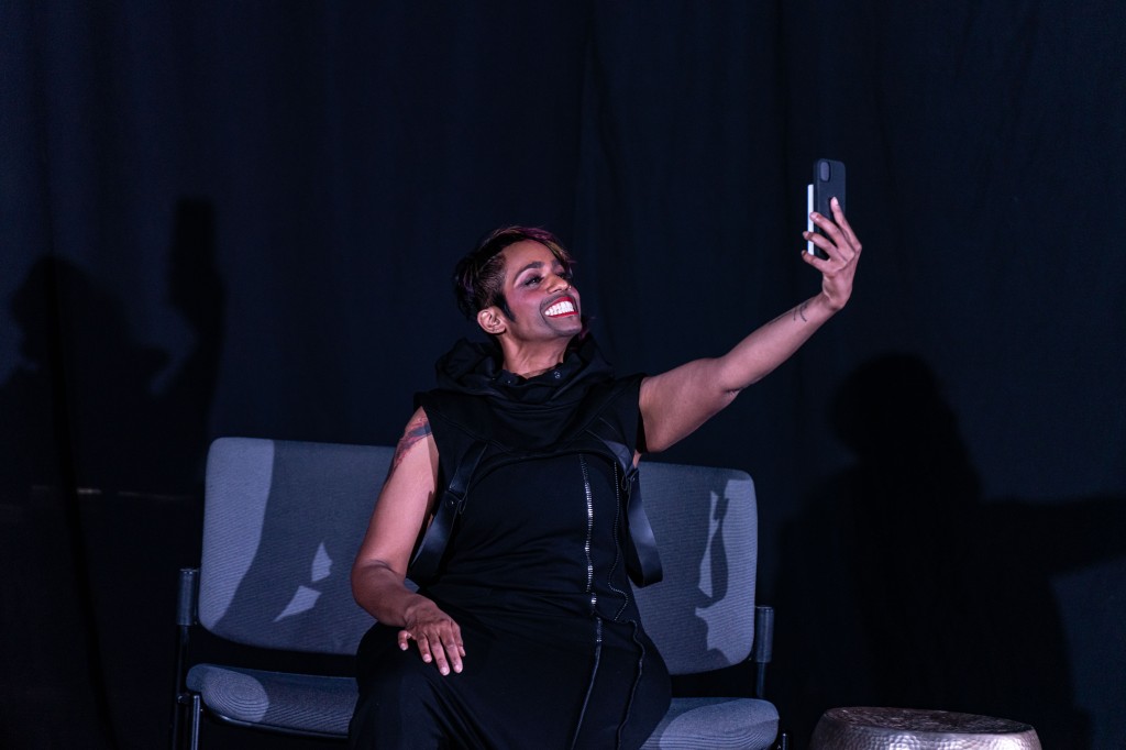 Photo caption: Dilsah is on a dark stage in black outfit, sitting on a chair looking into their smart phone smiling. 